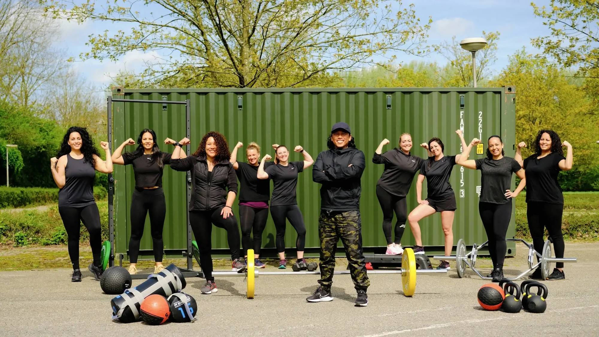Het STERK-team met gebalde vuisten voor de groene STERKBOX-container, met kettlebells, zandzakken en halters op de grond.