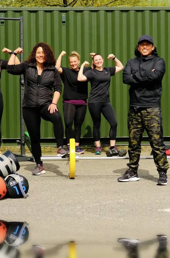Het STERK-team met gebalde vuisten voor de groene STERKBOX-container, met kettlebells, zandzakken en halters op de grond.