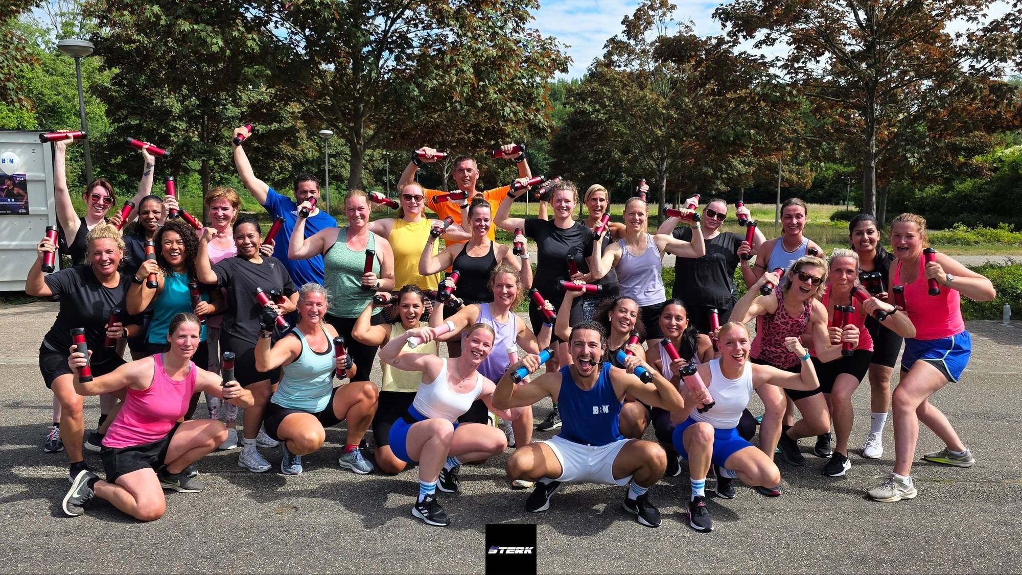 Een grote groep lachende sporters met dumbbells in de lucht na een buitentraining.