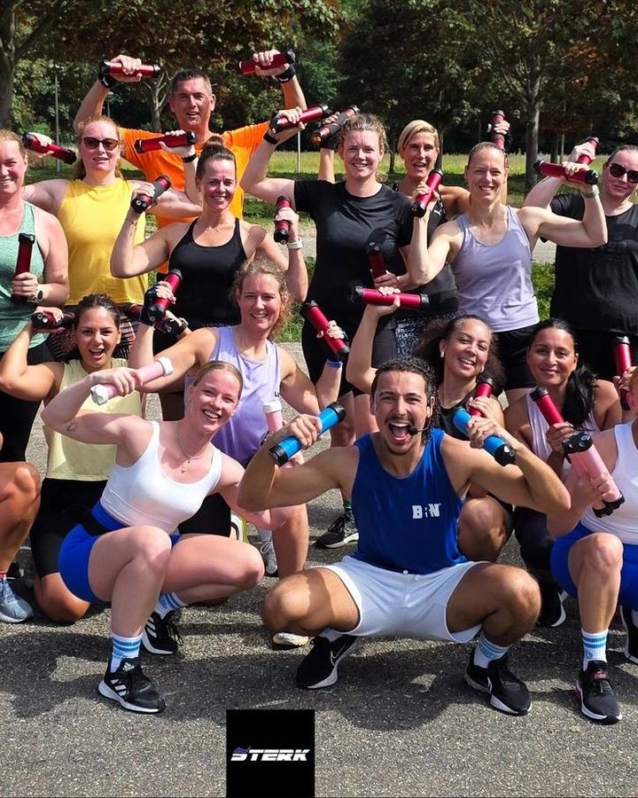 Een grote groep lachende sporters met dumbbells in de lucht na een buitentraining.