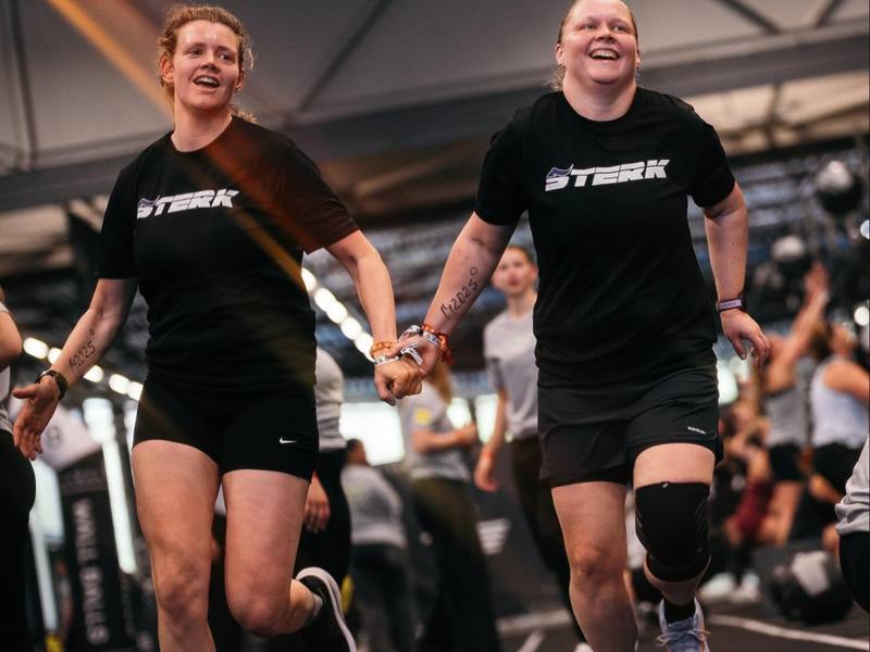 Twee sporters in STERK-shirts rennen hand in hand tijdens een HYROX-race.
