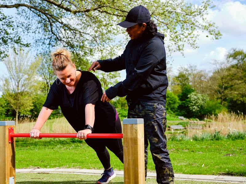 Een trainer begeleidt een sporter bij een oefening aan de stang in het park.