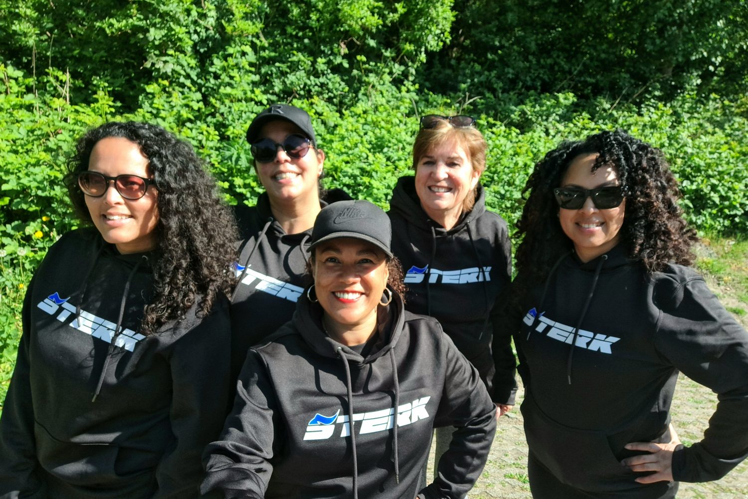 Vijf trainers van STERK in zwarte STERK-hoodies, lachend buiten in de zon.